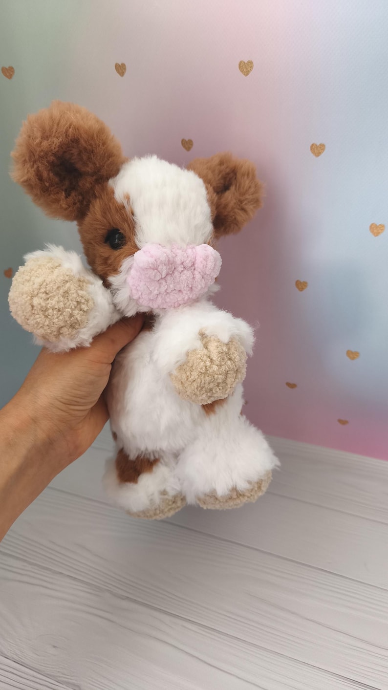 Pode incluir: Um brinquedo de pel&uacute;cia de vaca feito &agrave; m&atilde;o com pelo marrom e branco, orelhas grandes marrons e um focinho rosa. O brinquedo &eacute; segurado em uma m&atilde;o, mostrando sua textura macia e artesanato detalhado. O fundo apresenta um gradiente de rosa e azul com detalhes de cora&ccedil;&atilde;o dourado.