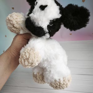 Pode incluir: Um brinquedo de pel&uacute;cia em forma de vaca com um padr&atilde;o preto e branco. A vaca tem orelhas grandes pretas, um rosto branco e um corpo branco fofo. Os cascos s&atilde;o bege. O brinquedo &eacute; segurado por uma m&atilde;o.