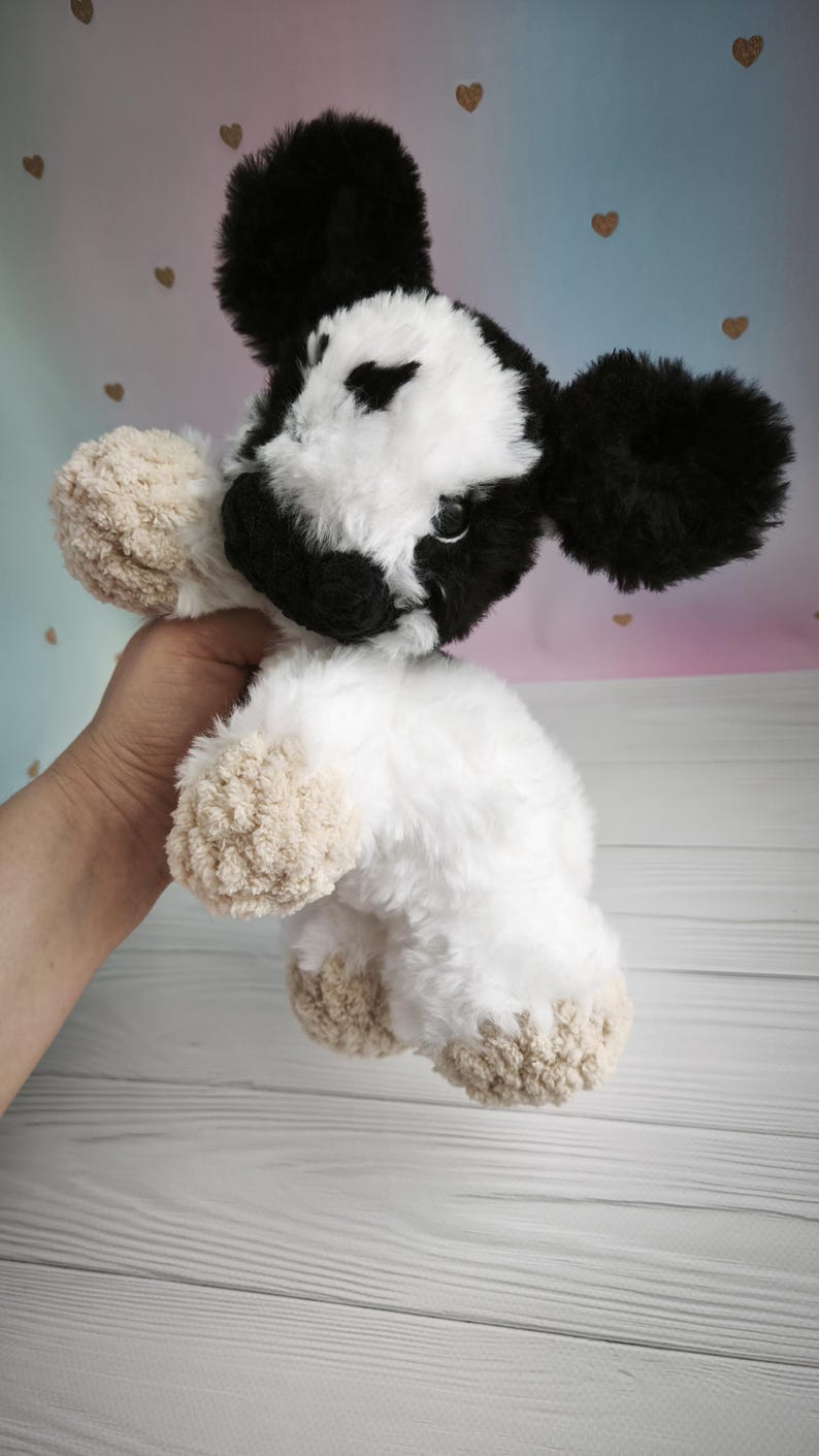 Pode incluir: Um brinquedo de pel&uacute;cia em forma de vaca com um padr&atilde;o preto e branco. A vaca tem orelhas grandes pretas, um rosto branco e um corpo branco fofo. Os cascos s&atilde;o bege. O brinquedo &eacute; segurado por uma m&atilde;o.