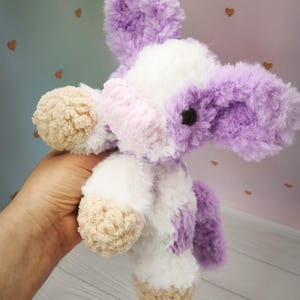 Pode incluir: Um brinquedo de pel&uacute;cia feito &agrave; m&atilde;o em forma de vaca. O brinquedo &eacute; branco com detalhes roxos e marrom claro. A vaca tem orelhas grandes e fofas e uma apar&ecirc;ncia macia e texturizada. O brinquedo &eacute; segurado na m&atilde;o de uma pessoa.