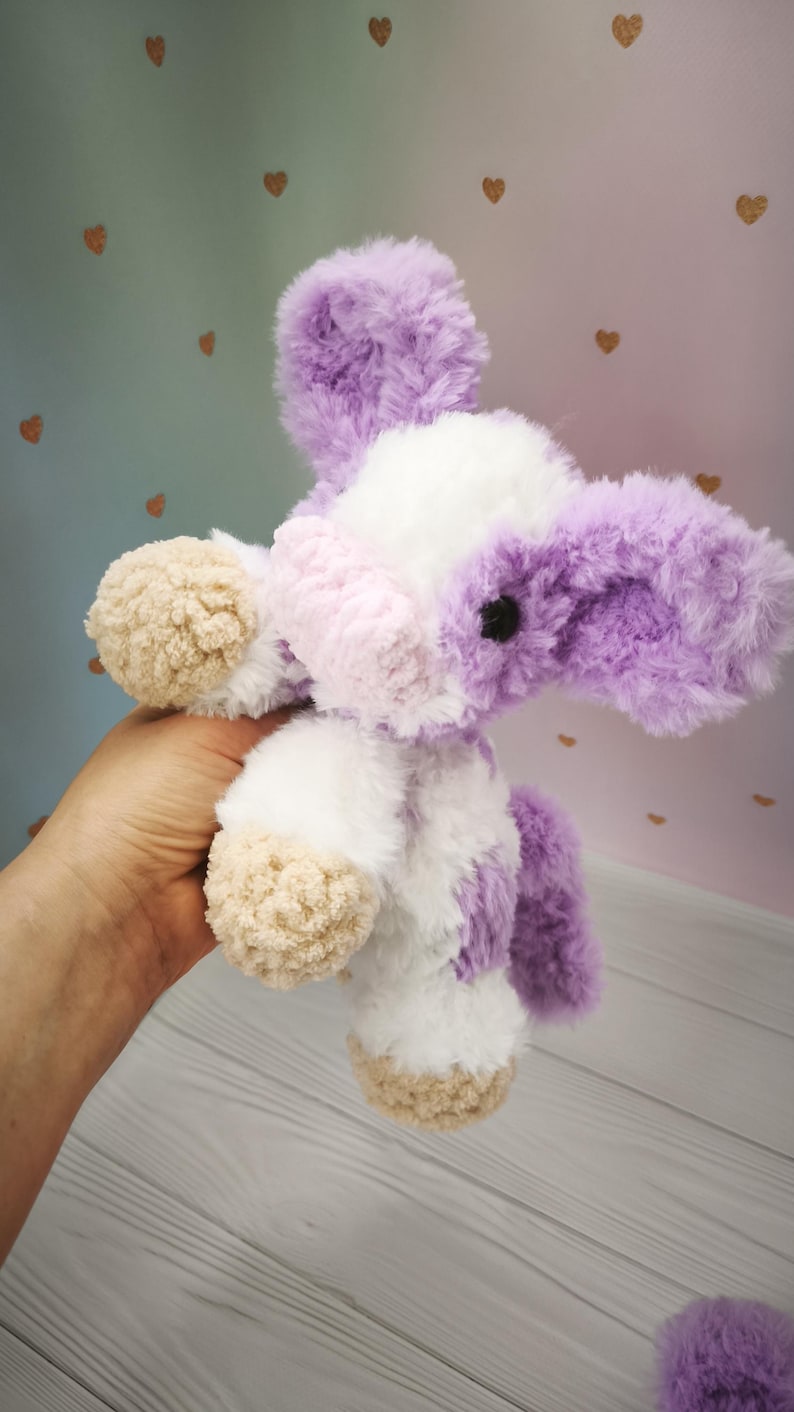 Pode incluir: Um brinquedo de pel&uacute;cia feito &agrave; m&atilde;o em forma de vaca. O brinquedo &eacute; branco com detalhes roxos e marrom claro. A vaca tem orelhas grandes e fofas e uma apar&ecirc;ncia macia e texturizada. O brinquedo &eacute; segurado na m&atilde;o de uma pessoa.