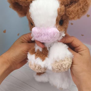 Pode incluir: Um brinquedo de pel&uacute;cia de vaca com pelo castanho e branco, orelhas grandes castanhas e um focinho rosa. O bicho de pel&uacute;cia &eacute; segurado em frente a um fundo macio e colorido.