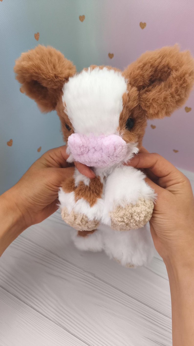 Pode incluir: Um brinquedo de pel&uacute;cia de vaca com pelo castanho e branco, orelhas grandes castanhas e um focinho rosa. O bicho de pel&uacute;cia &eacute; segurado em frente a um fundo macio e colorido.