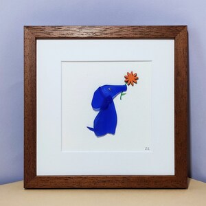 May include: A framed artwork featuring a blue glass dog sniffing an orange flower. The artwork is signed "ER".