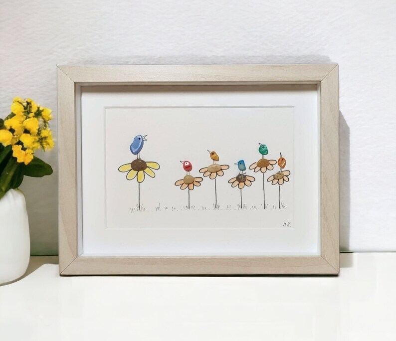 Op de afbeelding: Ingelijst kunstwerk met een kleurrijk kiezelsteenkunstontwerp van vogels die op gele bloemen zitten. De vogels zijn geschilderd in tinten blauw, groen, rood en geel. Het kunstwerk is gesigneerd met "2C".