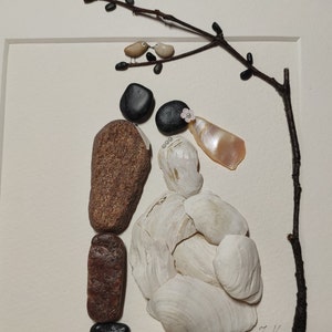 May include: A stone art piece depicting a bride and groom with a small branch above them. The bride is made of white seashells and the groom is made of brown and black stones. The branch has two small birds on it. The art piece is signed "J.K."