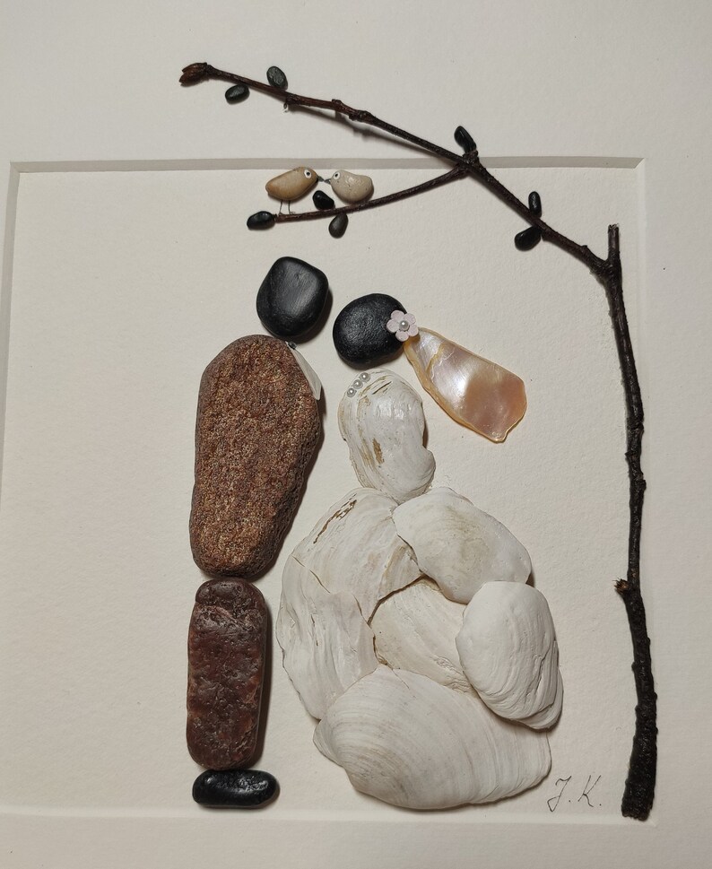May include: A stone art piece depicting a bride and groom with a small branch above them. The bride is made of white seashells and the groom is made of brown and black stones. The branch has two small birds on it. The art piece is signed "J.K."