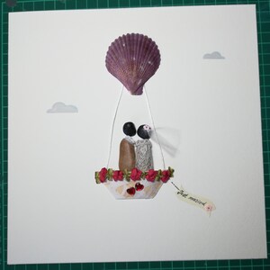 May include: A white card with a seashell hot air balloon carrying a bride and groom made of stones. The balloon is decorated with red flowers and the card says "Just married".