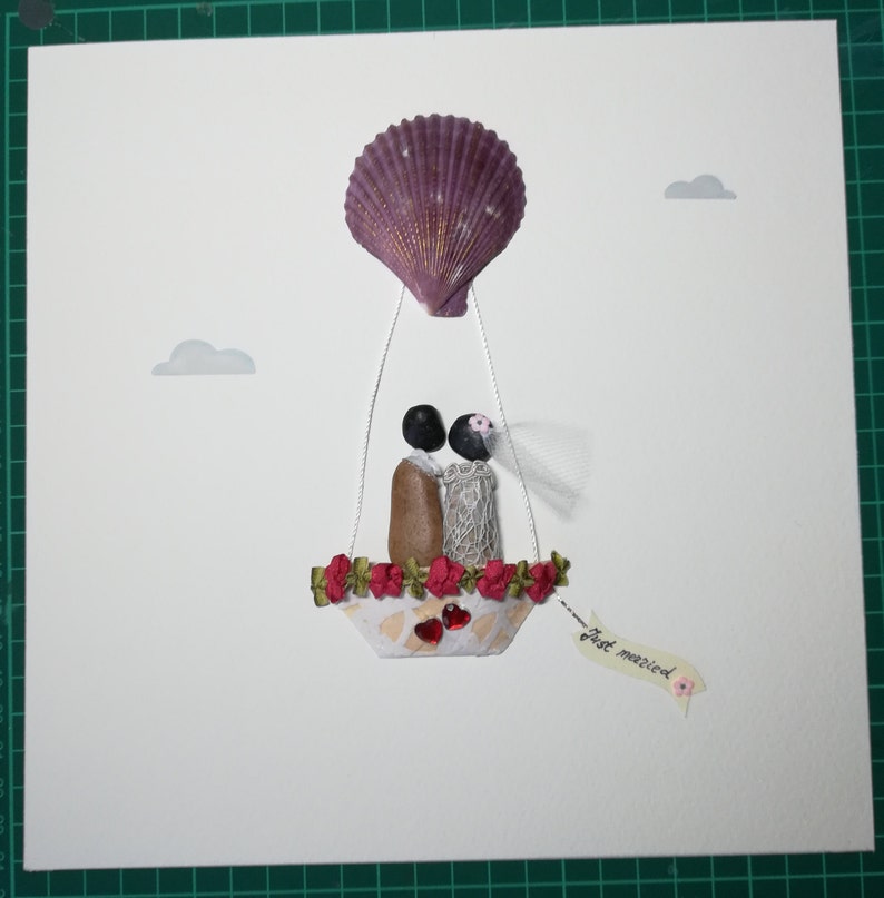 May include: A white card with a seashell hot air balloon carrying a bride and groom made of stones. The balloon is decorated with red flowers and the card says "Just married".