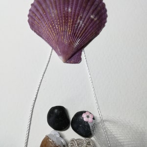 May include: A seashell wedding decoration with a purple seashell hanging from white string and two figures made of stones. The bride has a white veil and a pink flower. The groom has a brown stone body and a black stone head.