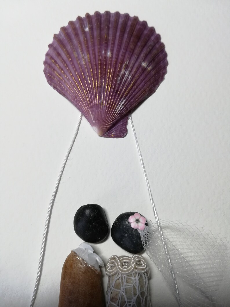 May include: A seashell wedding decoration with a purple seashell hanging from white string and two figures made of stones. The bride has a white veil and a pink flower. The groom has a brown stone body and a black stone head.