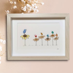 Op de afbeelding: Een ingelijst kunstwerk met een rij kleurrijke vogels die op gele bloemen zitten. De vogels zijn gemaakt van beschilderde stenen en de bloemen van kiezelsteentjes. Het kunstwerk is gesigneerd met "S.K."