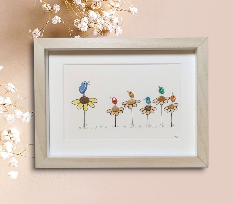 Op de afbeelding: Een ingelijst kunstwerk met een rij kleurrijke vogels die op gele bloemen zitten. De vogels zijn gemaakt van beschilderde stenen en de bloemen van kiezelsteentjes. Het kunstwerk is gesigneerd met "S.K."