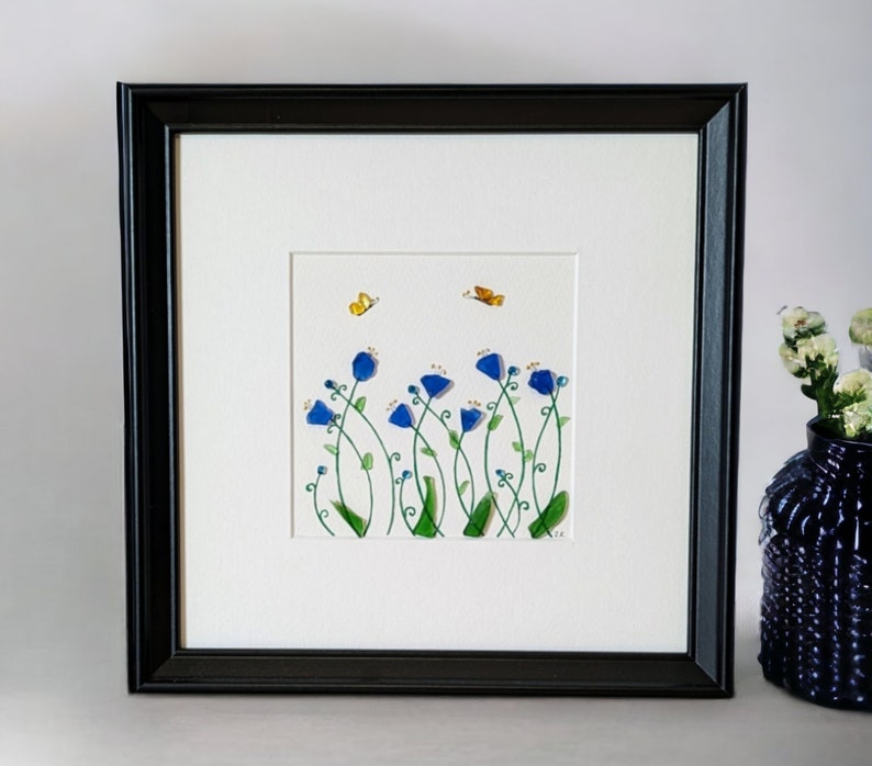 May include: A framed artwork featuring a watercolor painting of blue flowers with green stems and yellow butterflies on a white background.