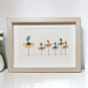 Op de afbeelding: Een ingelijst kunstwerk met een rij kleurrijke vogels die op gele bloemen zitten. De vogels zijn gemaakt van beschilderde stenen en de bloemen zijn geschilderd op een witte achtergrond. Het kunstwerk is gesigneerd met "JC".