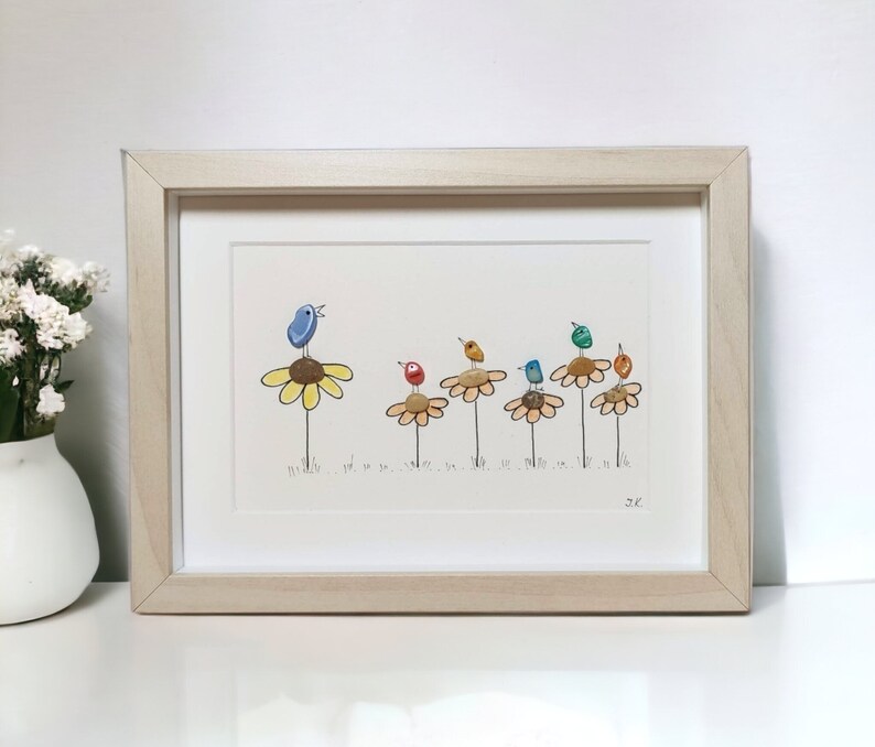 Op de afbeelding: Een ingelijst kunstwerk met een rij kleurrijke vogels die op gele bloemen zitten. De vogels zijn gemaakt van beschilderde stenen en de bloemen zijn geschilderd op een witte achtergrond. Het kunstwerk is gesigneerd met "JC".