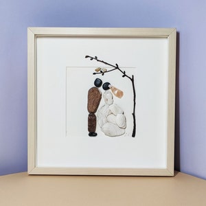 May include: A framed artwork made of small stones depicting a bride and groom standing under a tree branch. The bride is made of white stones and the groom is made of brown and black stones.