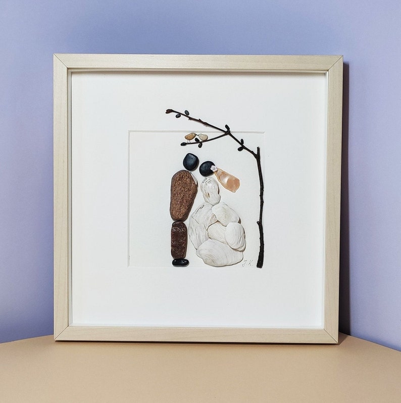 May include: A framed artwork made of small stones depicting a bride and groom standing under a tree branch. The bride is made of white stones and the groom is made of brown and black stones.
