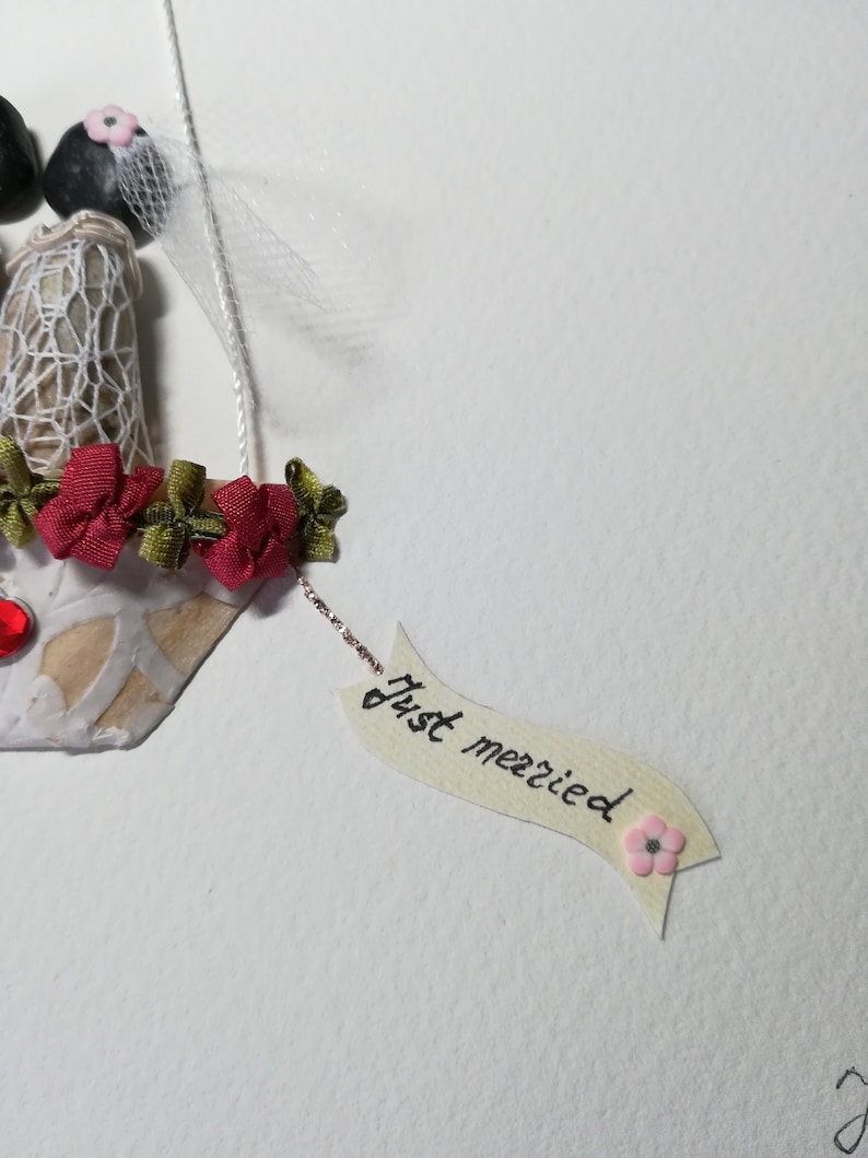 May include: A handmade wedding card with a couple made of stones, a white veil, and red flowers. The card has a banner that says "Just married" with a pink flower.