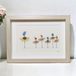 Op de afbeelding: Een ingelijst kunstwerk met een rij kleurrijke vogels die op gele bloemen zitten. De vogels zijn gemaakt van beschilderde stenen en de bloemen van beschilderde kiezelsteentjes. Het kunstwerk is rechtsonder getekend met "JK".
