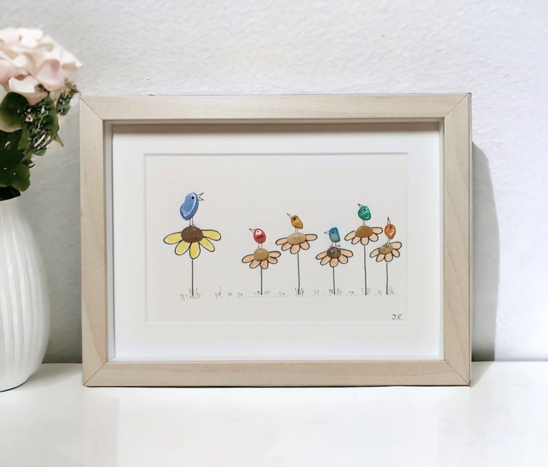Op de afbeelding: Een ingelijst kunstwerk met een rij kleurrijke vogels die op gele bloemen zitten. De vogels zijn gemaakt van beschilderde stenen en de bloemen van beschilderde kiezelsteentjes. Het kunstwerk is rechtsonder getekend met "JK".