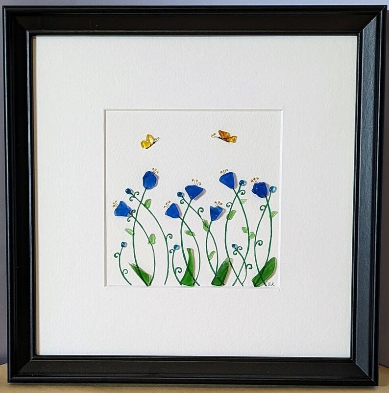 May include: A framed artwork featuring a blue and green glass mosaic of flowers and butterflies on a white background. The artwork is signed "SK" in the bottom right corner.