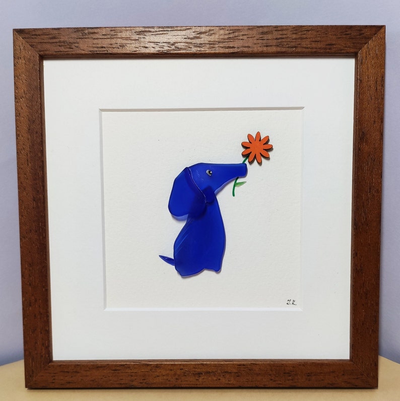 May include: A framed artwork featuring a blue glass dog sniffing a red flower. The artwork is on a white background and has a brown frame.