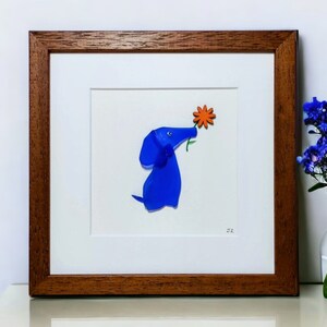May include: A framed artwork featuring a blue glass dog with an orange flower in its mouth. The artwork is set against a white background and has a brown wooden frame.