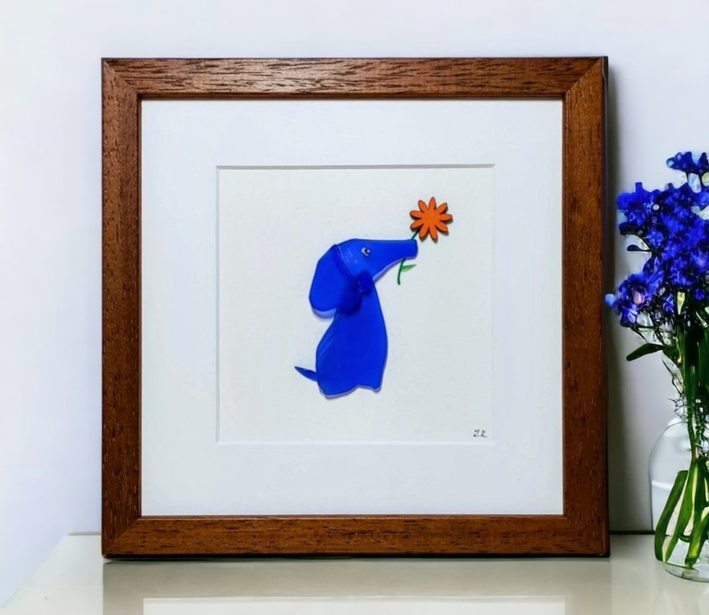 May include: A framed artwork featuring a blue glass dog with an orange flower in its mouth. The artwork is set against a white background and has a brown wooden frame.