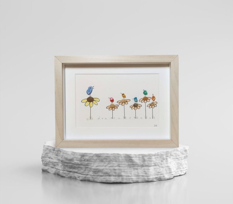 Op de afbeelding: Een ingelijst kunstwerk met een grillige illustratie van kleurrijke vogels die op bloemstelen zitten. De vogels zijn gemaakt van beschilderde kiezelsteentjes en de bloemen zijn in een eenvoudige stijl getekend. Het kunstwerk is gesigneerd met "2C".