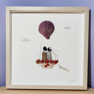 May include: A framed artwork featuring a seashell hot air balloon with a bride and groom inside. The balloon is decorated with red flowers and the couple is holding hands. The artwork has a white background and a wooden frame. The text "Just married" is written on a tag hanging from the balloon.