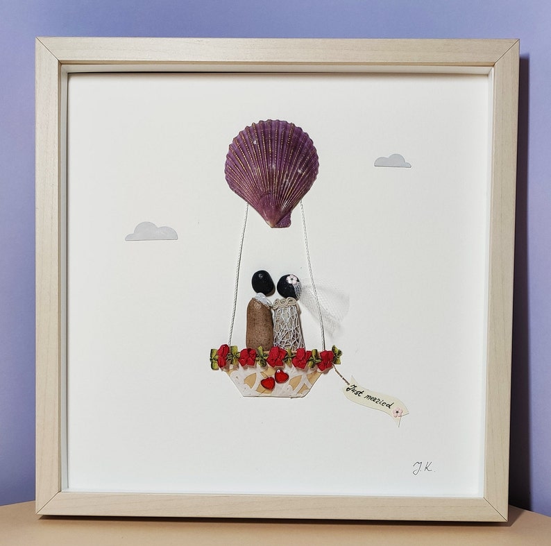 May include: A framed artwork featuring a seashell hot air balloon with a bride and groom inside. The balloon is decorated with red flowers and the couple is holding hands. The artwork has a white background and a wooden frame. The text "Just married" is written on a tag hanging from the balloon.