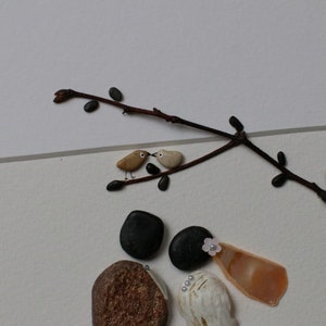 May include: A framed pebble art piece depicting a couple, a bride and groom, with a small branch with two birds perched on it. The bride is wearing a white shell and a pink flower. The groom is wearing a black stone hat. The background is white.