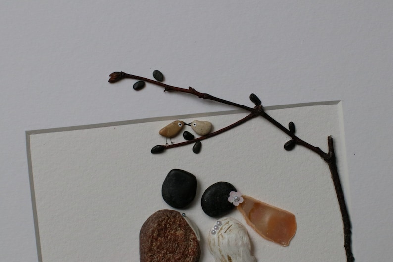 May include: A framed pebble art piece depicting a couple, a bride and groom, with a small branch with two birds perched on it. The bride is wearing a white shell and a pink flower. The groom is wearing a black stone hat. The background is white.