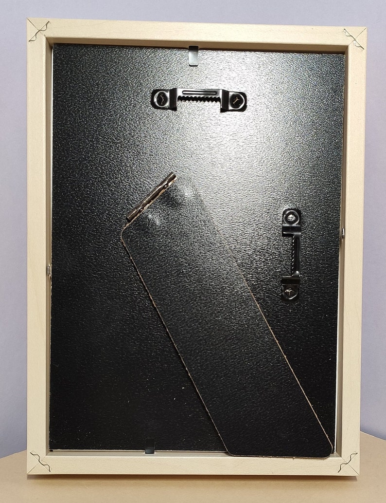Op de afbeelding: Een licht houten fotolijst met een zwarte achterkant en een zwarte ezelstandaard. De lijst heeft twee metalen ophanghaken aan de achterkant.