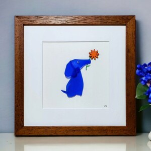 May include: A framed artwork featuring a blue glass dog sniffing an orange flower. The artwork is set against a white background and has a brown wooden frame.