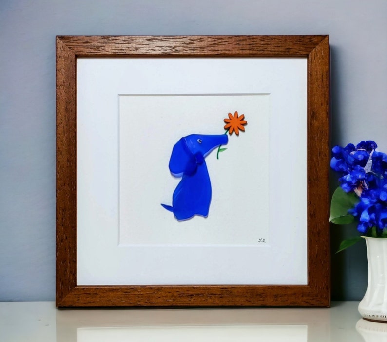 May include: A framed artwork featuring a blue glass dog sniffing an orange flower. The artwork is set against a white background and has a brown wooden frame.