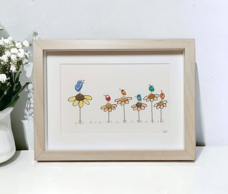 Op de afbeelding: Een ingelijst kunstwerk met een rij kleurrijke vogels die op gele bloemen zitten. De vogels zijn gemaakt van beschilderde stenen en de bloemen van beschilderde kiezelsteentjes. Het kunstwerk is gesigneerd met "JC".