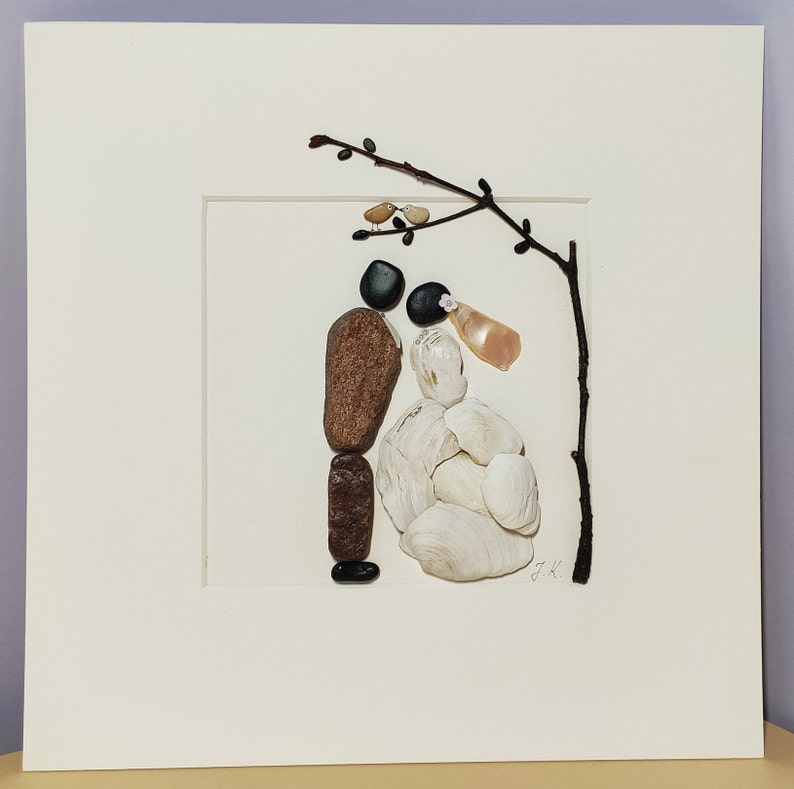 May include: A framed artwork made with stones and shells depicting a bride and groom standing under a branch with two birds perched on it. The artwork is signed "J.K."