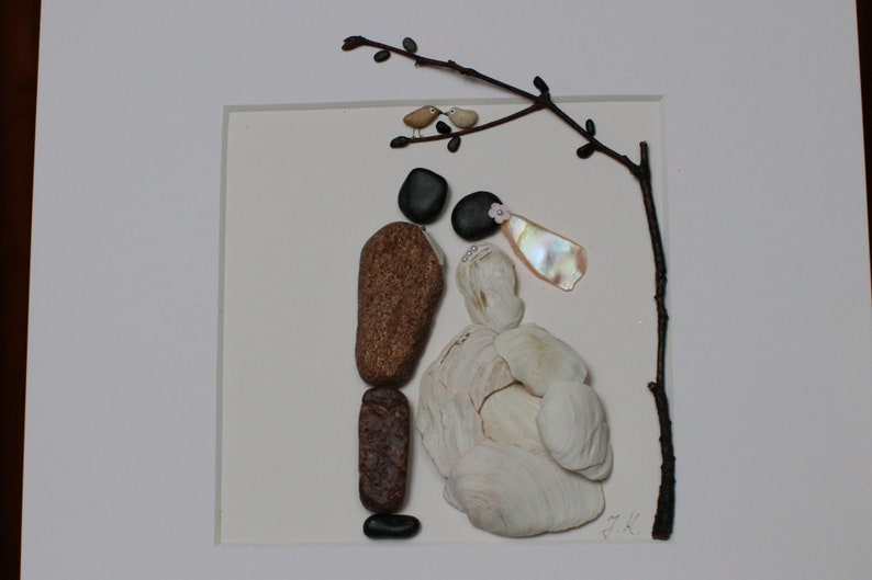 May include: A framed artwork made with stones and shells depicting a bride and groom standing under a branch with two birds perched on it. The bride is wearing a white dress made of shells and the groom is wearing a brown suit made of stones. The artwork is signed "D.K."