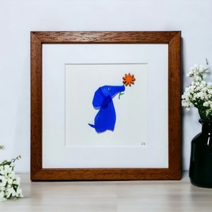 May include: A framed artwork featuring a blue glass dog sniffing an orange flower. The artwork is set against a white background and framed in a brown wood frame.