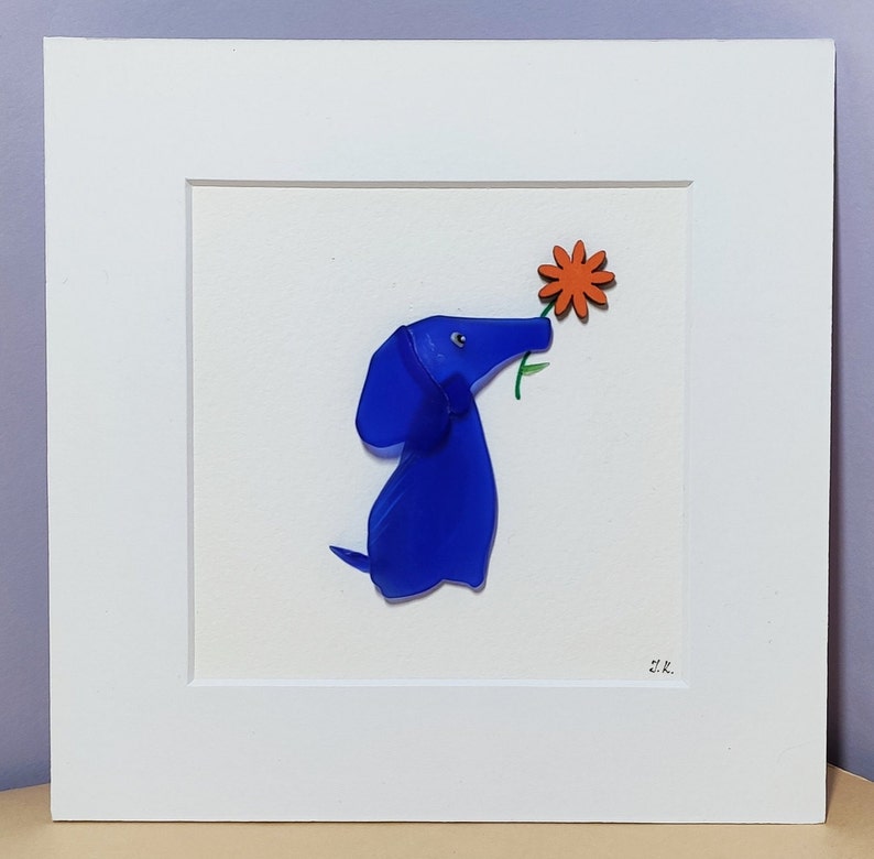 May include: A blue glass dog sculpture with a flower in its mouth. The dog is standing on a white background. The flower is orange and has six petals. The sculpture is framed in a white frame.