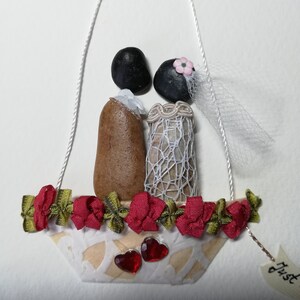 May include: A wedding-themed pebble art piece featuring a bride and groom in a white boat with red and green ribbon trim. The boat is decorated with two red hearts and the text "Just" is visible on a tag.