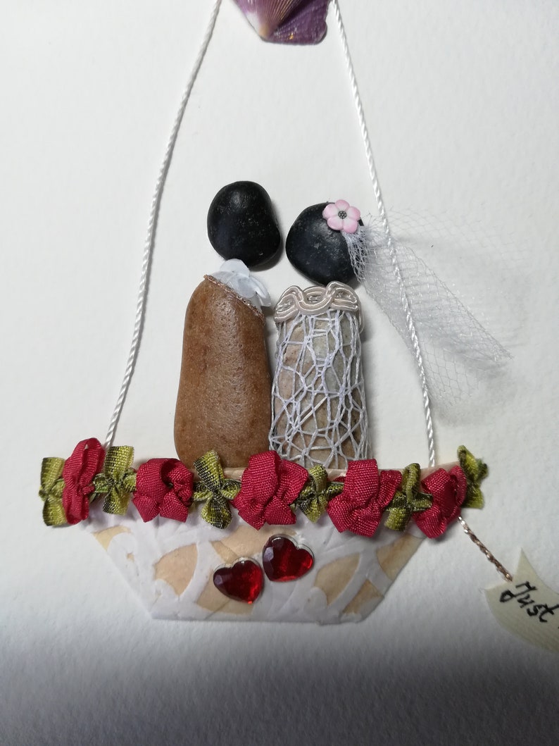 May include: A wedding-themed pebble art piece featuring a bride and groom in a white boat with red and green ribbon trim. The boat is decorated with two red hearts and the text "Just" is visible on a tag.