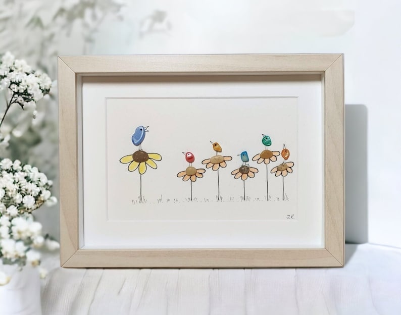 Op de afbeelding: Ingelijst kunstwerk met een kiezelkunstontwerp van kleurrijke vogels die op gele bloemen zitten. De vogels zijn gemaakt van beschilderde stenen en de bloemen van kiezels. Het kunstwerk is gesigneerd met "JC".
