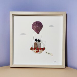 May include: A framed artwork featuring a seashell hot air balloon with a couple inside. The balloon is decorated with red flowers and the couple is holding hands. The artwork has a white background and a wooden frame. The text "Just married" is written on a tag hanging from the balloon.