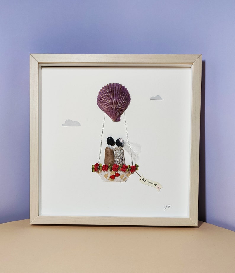 May include: A framed artwork featuring a seashell hot air balloon with a couple inside. The balloon is decorated with red flowers and the couple is holding hands. The artwork has a white background and a wooden frame. The text "Just married" is written on a tag hanging from the balloon.