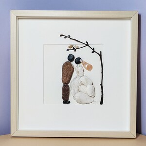 May include: A framed artwork depicting a couple made of stones, standing under a twig with small white flowers. The bride is made of white stones and the groom is made of brown and black stones.