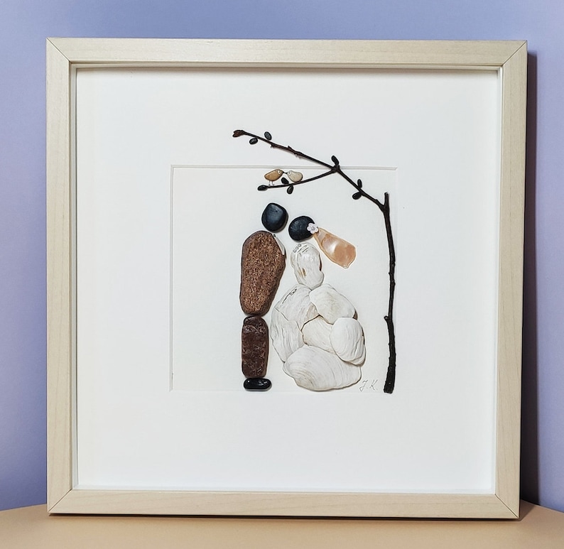 May include: A framed artwork depicting a couple made of stones, standing under a twig with small white flowers. The bride is made of white stones and the groom is made of brown and black stones.