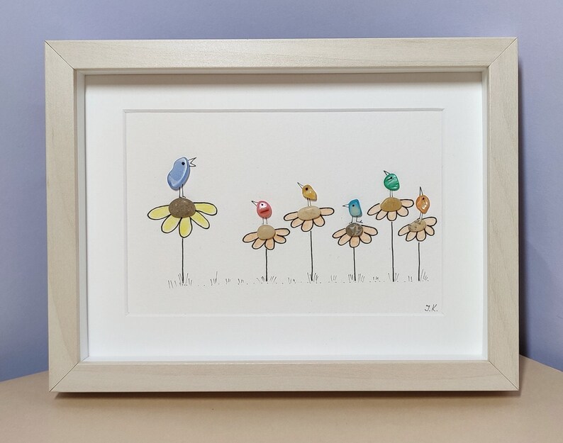 Op de afbeelding: Een ingelijst kunstwerk met een blauwe vogel die zingt op een gele bloem, met vijf andere kleurrijke vogels die op gele bloemen zitten. Het kunstwerk is gemaakt van kiezelsteentjes en is gesigneerd met "J.K."